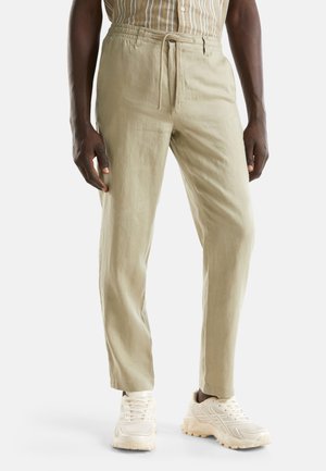 Hombre con pantalones beige con cordón, zapatillas blancas gruesas y una camisa de rayas metida por dentro, de pie con los brazos relajados a los lados.