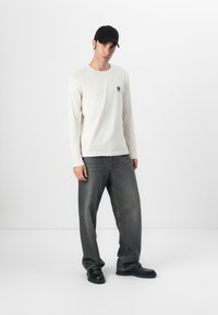 Long-sleeve cream t-shirt with a chest logo, paired with loose, faded gray denim jeans and black shoes. Model wears a black cap.