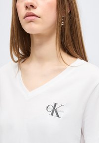 Νεαρή γυναίκα που φοράει λευκό T-shirt Calvin Klein με V-λαιμόκοψη και λογότυπο στο αριστερό στήθος και μαργαριταρένια σκουλαρίκια, εμφανίζεται από τον λαιμό έως το μέσο του κορμού.