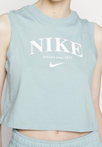 Světlomodrý bezrukávový crop top vyrobený z měkké bavlny, s bílým potiskem loga Nike a textem "Athletic dept. 1972" pod ním.