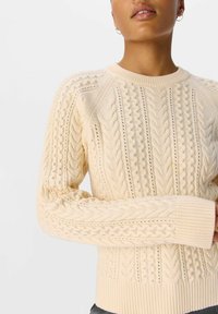 Krämsfärgad stickad tröja med kabelmönster, ribbad ärmslut och hem, med en strukturerad mönstring genom hela plagget och rund halsringning.