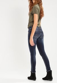 Jeans ajustados en denim azul oscuro con una textura desvanecida y detalles de costura sutiles, combinados con una camiseta verde oliva y botas negras hasta el tobillo.