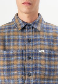 Blue and orange plaid flannel shirt with a buttoned collar, black buttons, and a small Wrangler logo stitched on the chest.