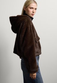 Chaqueta corta de pelaje sintético marrón con capucha, que incluye un bolsillo frontal y puños acanalados. Combinada con unos vaqueros de talle alto azules.