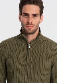 Groene gebreide pullover met een gestructureerd patroon, ritsdetail bij de kraag en ribgebreide hals. Strakke pasvorm en middelgewicht stof.