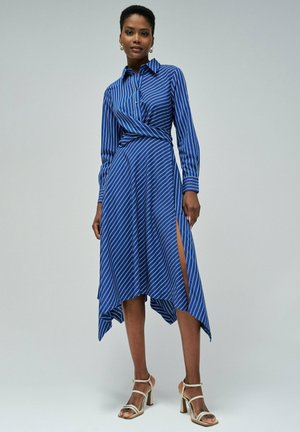 Vestido envolvente a rayas azules con mangas largas, dobladillo asimétrico, abertura lateral y cuello de camisa; acompañado de tacones metálicos con tiras.