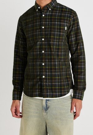 Uomo che indossa una camicia a quadri verde scuro, nera e gialla con taschino sul petto, abbinata a jeans blu chiaro.