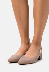 Dei tacchi slingback beige in suede con punta affusolata, tacco cubano basso e dettagli con fibbia circolare alla caviglia. Texture liscia e design elegante.
