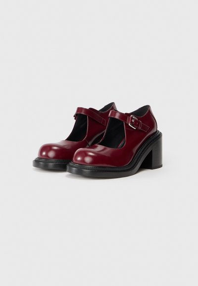 Chaussures T-bar en cuir bordeaux avec un large talon noir et une sangle réglable. Elles disposent d'un bout rond et d'un design découpé pour la ventilation.