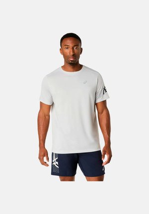 Athletischer Mann steht und trägt ein weißes Kurzarmshirt mit Logo sowie marineblaue Shorts mit weißen grafischen Mustern am linken Bein.