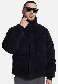 Black corduroy puffer jacket, featuring a high collar, diagonal front pockets, and ribbed cuffs. Smooth texture with quilted pattern.