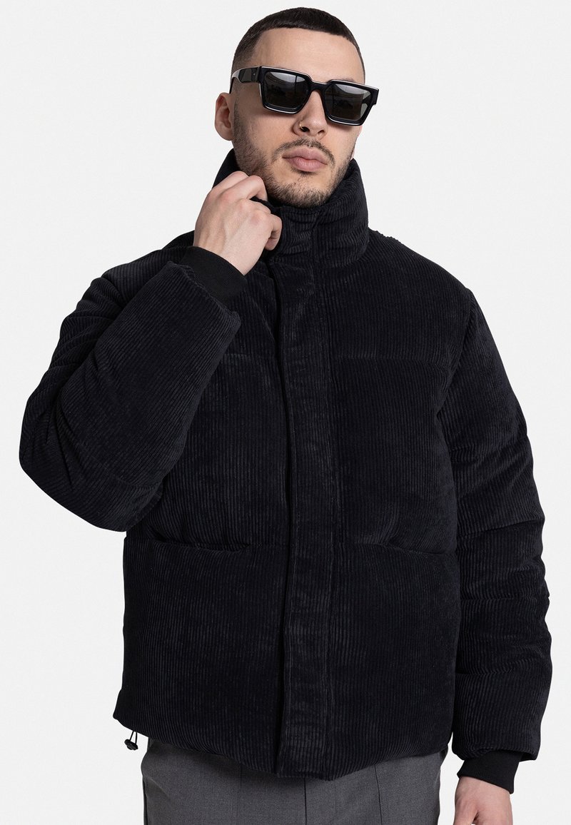 Black corduroy puffer jacket, featuring a high collar, diagonal front pockets, and ribbed cuffs. Smooth texture with quilted pattern.