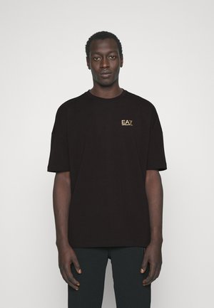 T-shirt in cotone nero con maniche corte, con un logo ricamato in oro sul petto a sinistra. Il modello indossa pantaloni scuri, in piedi contro uno sfondo neutro.