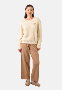 Maglione cremoso e morbido con un accento dorato a forma di cuore, abbinato a pantaloni larghi brillanti di colore marrone chiaro e scarpe slip-on beige.