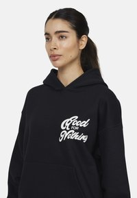 Felpa nera con cappuccio realizzata in tessuto morbido, presenta una tasca frontale e una grafica testuale bianca con la scritta "Good for Nothing". Cappuccio regolabile.