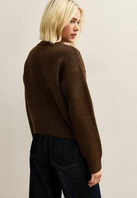 Brązowy sweter dzianinowy o krótkim kroju i długich rękawach, w połączeniu z ciemnymi dżinsami. Miękka faktura i szeroki, luźny krój.