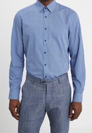 Mann trägt ein hellblaues, gemustertes Hemd, das in grau karierte Hosen gesteckt ist, vor einfarbigem Hintergrund.