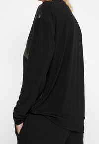 Svart långärmad topp med avslappnad passform, mjuk textur och draperad design. Inkluderar en kontrasterande detaljpanel på axlarna.