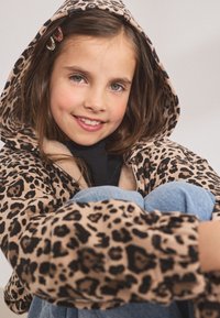 Leopardikuvioitu vetoketjullinen huppari, jossa on beige pohjaväri, mustia laikkuja, vuorattu sisäpuoli ja iso huppu. Vaaleansiniset farkut ja hiusklipsit tuovat yksityiskohtia.