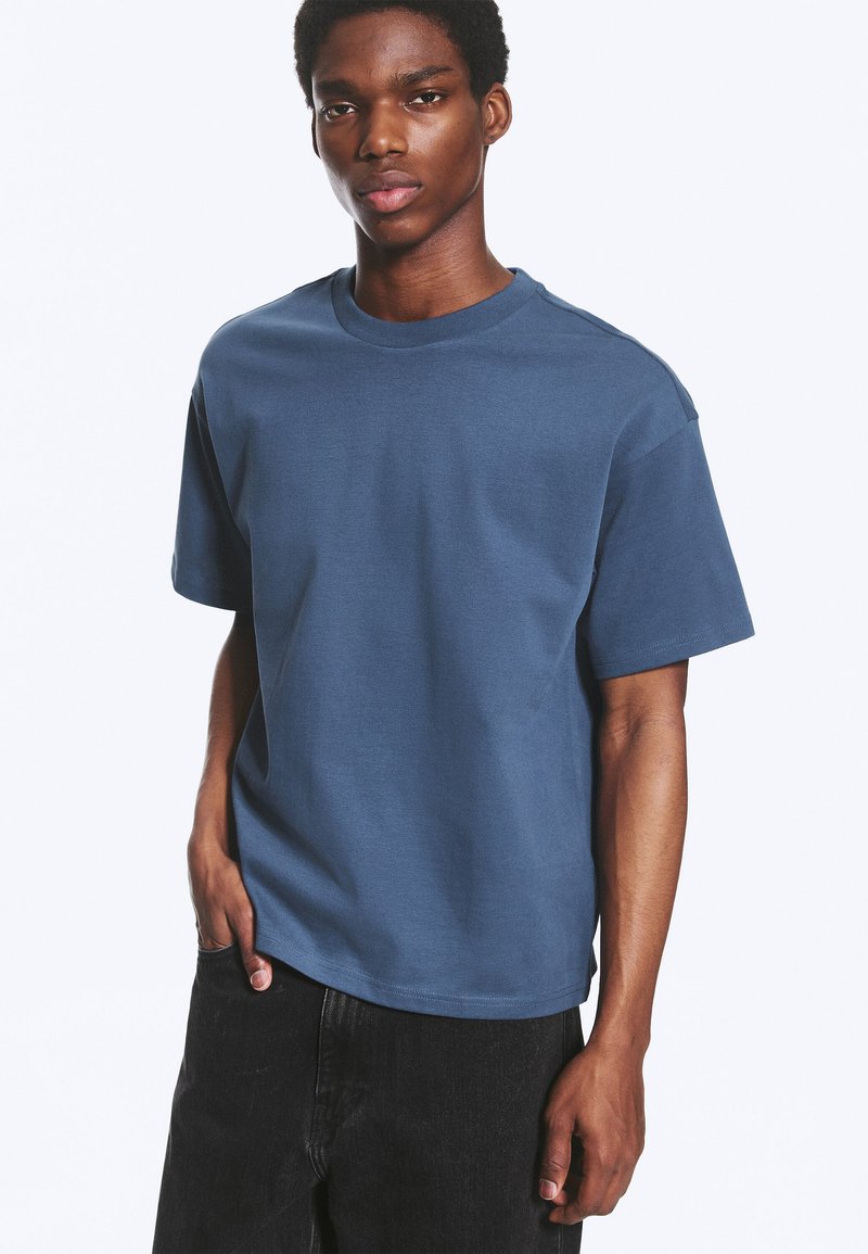 Jeune homme aux cheveux courts et bouclés portant un t-shirt bleu uni à manches courtes et un pantalon noir, debout avec une main dans la poche.