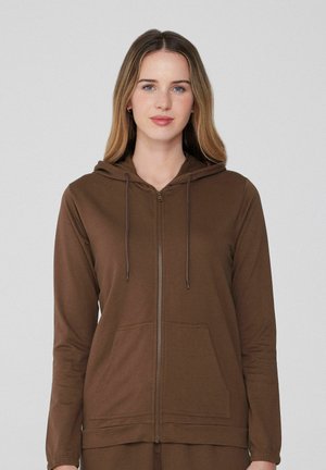 Junge Frau mit hellbraunen Haaren, die einen braunen Reißverschluss-Hoodie mit Fronttaschen und Kordeln trägt, steht vor einem schlichten Hintergrund.
