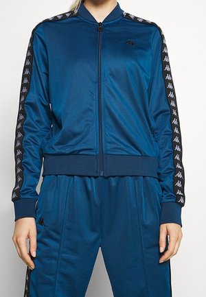 Femme portant un survêtement bleu à fermeture éclair intégrale avec des bandes noires et blanches à logo le long des manches et des jambes, debout devant un fond uni.