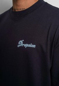 Marineblå bomulds T-shirt med rund hals; har tekstlogoen "Dropsize" i lyseblå på venstre brystområde. Glat tekstur.