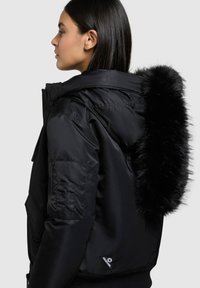 Geacă neagră matlasată cu glugă căptușită cu blană, buzunar lateral cu fermoar și tiv striat. Finisaj neted, mat, cu siluetă curbată.