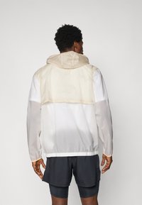 Giacca leggera con cappuccio in beige e bianco. Caratterizzata da polsi elasticizzati, orlo regolato e uno strato superiore trasparente. Dettaglio del logo riflettente.