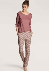 Lange mauve top met lange mouwen, gecombineerd met een broek met een patroon in een geometrisch ontwerp in roze en groen, voorzien van zijzakken en taps toelopende pijpen.