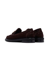 Mocasines de ante marrón con un bajo tacón negro, que presentan costuras decorativas y una pequeña correa en la parte delantera. La textura suave mejora su diseño.