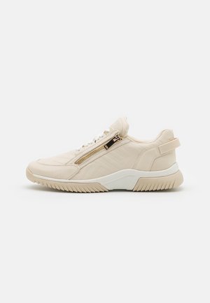 Beige lage sneaker met zijrits, veters aan de voorkant, getextureerde zool en trekhaak bij de hiel op een effen lichte achtergrond.