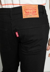 Detailní záběr na zadní kapsu černých džín Levi's, ukazující koženou značkovou nášivku se velikostí a červenou štítkovou značkou Levi's.