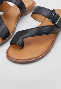 Sandalias de cuero negro con un diseño de tira ancha, un lazo para el dedo del pie y una plantilla de cuero marrón. Presentan una costura decorativa blanca alrededor de la suela.