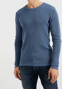 Blå långärmad stickad tröja med texturerad väv, rundad halsringning och åtsittande design. Buren med mörka denimjeans som visar en subtil logotypdetalj.