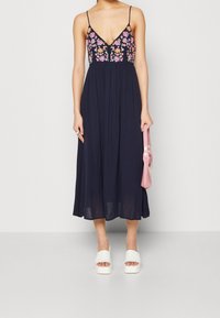 Námořnické maxi šaty s květinovou výšivkou na korzetu, tenkými ramínky, splývavou sukní, v kombinaci s bílými sandály na silném podpatku a růžovou kabelkou.