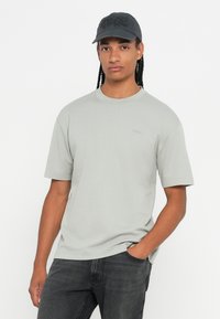 Νεαρός άνδρας με μακριές πλεξούδες, φορώντας μαύρο καπέλο, ανοιχτό γκρι υπερμεγέθη μπλούζα και σκούρες τζιν παντελόνες, που στέκεται με το ένα χέρι στην τσέπη.