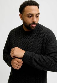 Black cable-knit sweater with a textured pattern, ribbed collar, and cuffs. Model is shown adjusting his sleeves.