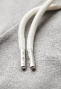 Jasnoszara bluza z kapturem z białymi bawełnianymi sznurkami. Metalowe końcówki sznurków z wygrawerowanym napisem "SUPERDRY" dodają szczegółu do projektu. Teksturowane tło.