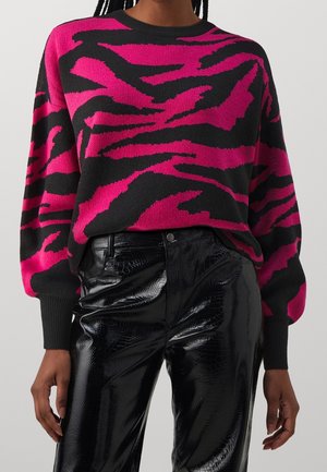 Persoon draagt een roze en zwart abstract patroon trui gecombineerd met zwarte, glanzende, hoog getailleerde nep leren broek met krokodillenstructuur.