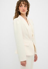 Blazer su misura color crema con maniche lunghe e strutturate e rever a punte. Presenta una chiusura a un bottone e una silhouette aderente.