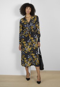 Robe fleurie noire avec des motifs jaunes et gris, manches longues, taille nouée et fente à l'avant, associée à un sac à main noir.