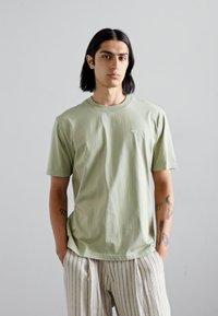 Tricou din bumbac de culoare verde deschis, cu decolteu rotund și mâneci scurte, asociat cu pantaloni bej cu dungi. Prezentă un mic logo pe piept.