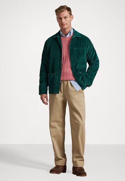 Men's green corduroy jacket with front pockets, layered over a pink cable-knit sweater and blue button-up shirt, paired with beige trousers.