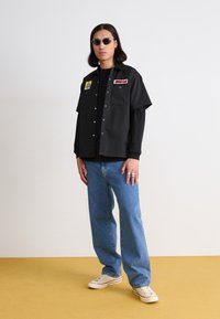 Črna kratka majica s patkami, črna majica pod njo, modri jeans in bele superge, dodatno opremljena s krožnimi sončnimi očali.