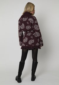 Mujer con botas negras y un vestido de punto con patrón paisley en morado oscuro y con flecos en el dobladillo, de pie mirando hacia atrás contra un fondo simple.