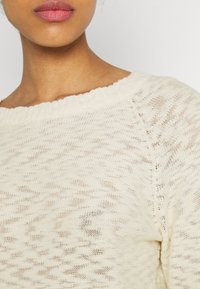 Ljus beige stickad tröja med bred halsringning, med ett strukturerat mönster och en ribbad fåll. Tyget verkar mjukt och luftigt.