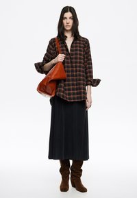 Camisa de gran tamaño a cuadros en negro y naranja, falda larga oscura, botas altas de ante marrón y bolso de cuero naranja. Diseño simple y moderno.