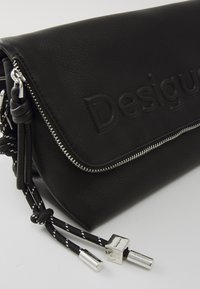 Crna kožna torbica s reljefnim logotipom, srebrnim patentnim zatvaračem i pletenim trakama s metalnim detaljima. Tekstuirana površina s elegantnim dizajnom.