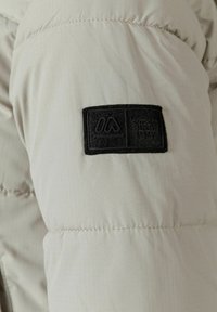 Veste rembourrée gris clair montrant un tissu texturé et des détails de coutures, avec une étiquette logo rectangulaire noire portant la mention "Outdoor Since 1997".